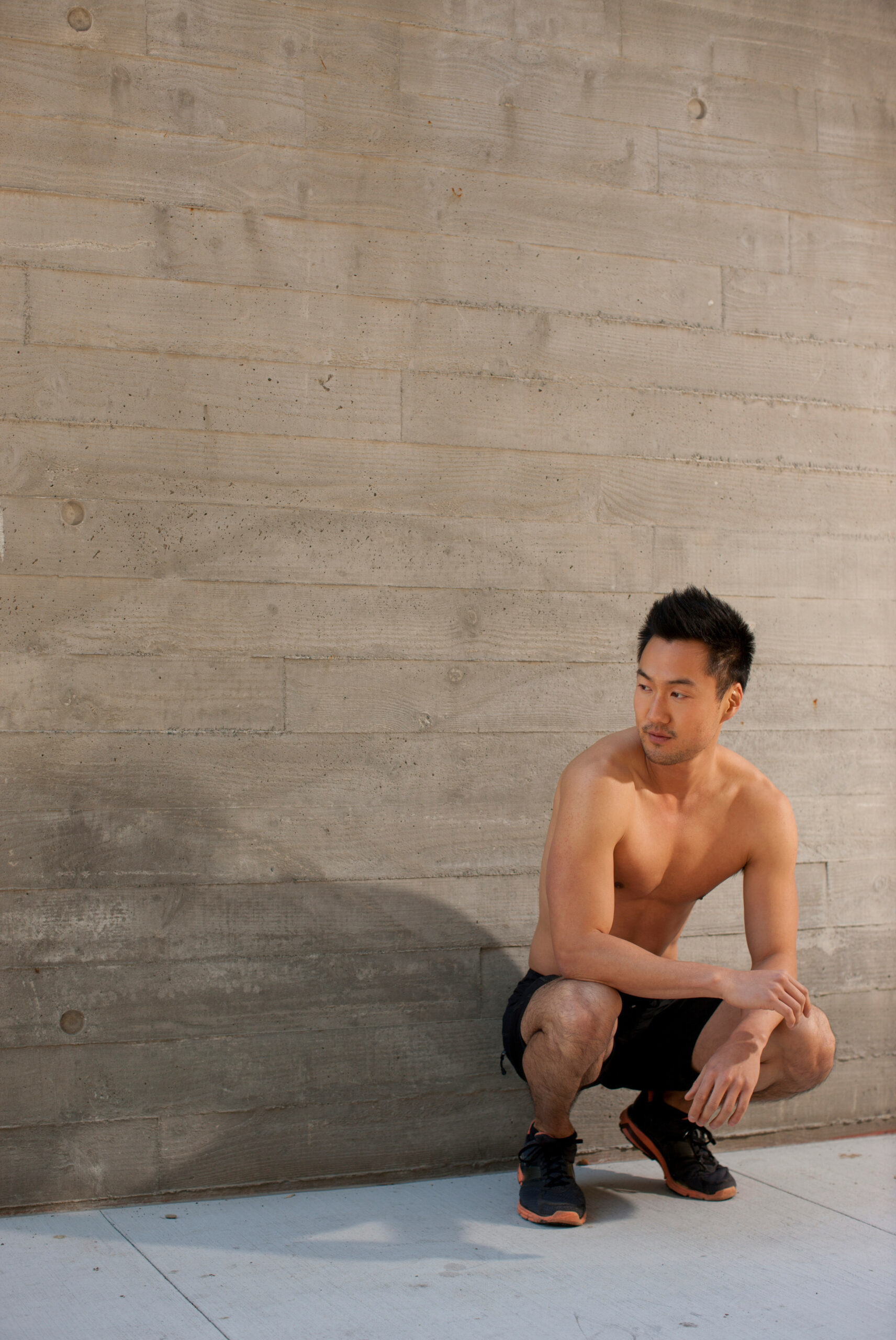 Shirtless man squatting against concrete wall outdoors.