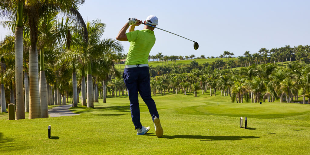 Golfer swinging on lush green course with palm trees.
