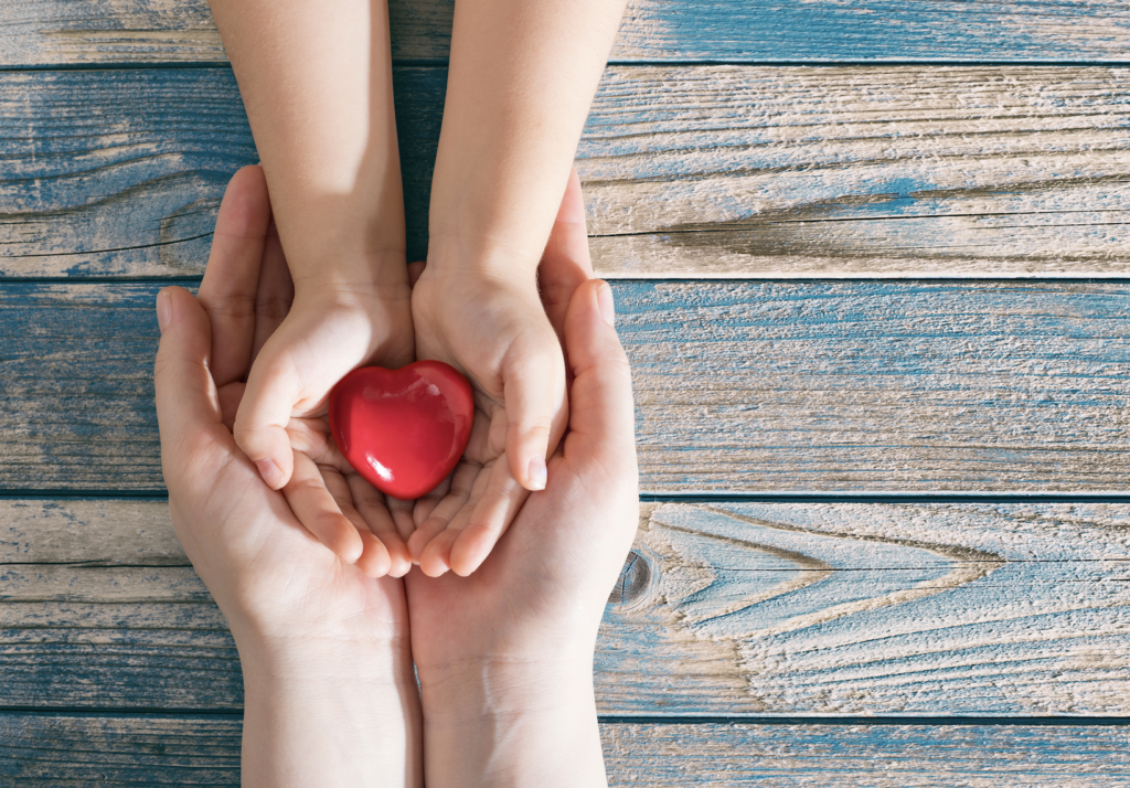 Hands holding a red heart on wooden background.